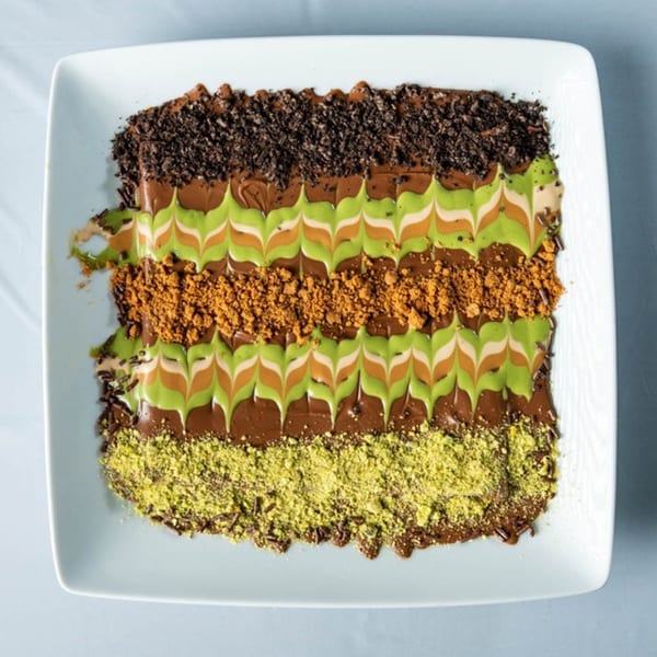 Ranoush dessert with chocolate, pistachio, and cookie crumbles in decorative stripes on a white plate.