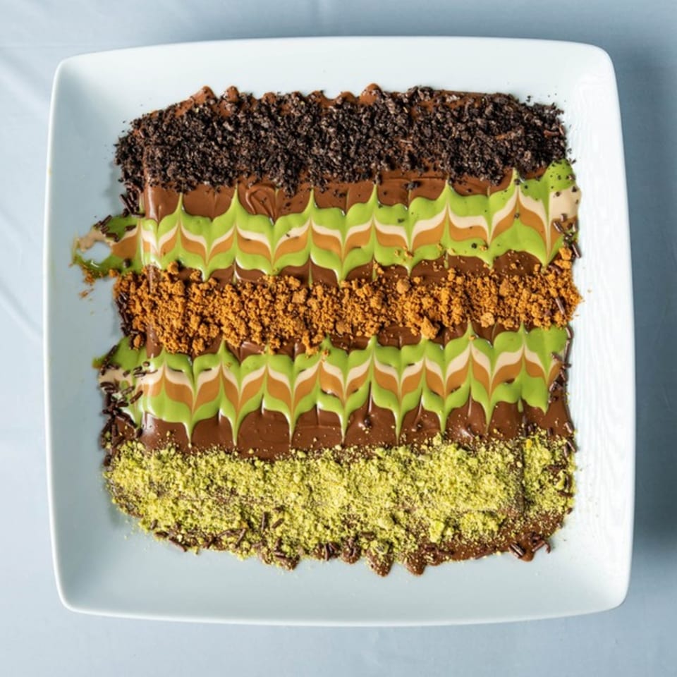 Ranoush dessert with chocolate, pistachio, and cookie crumbles in decorative stripes on a white plate.