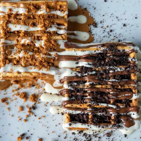 Two Ranoush waffles, one with cookie crumbs and the other with Lotus Biscoff crumbs, drizzled with white and milk chocolate.